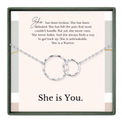 The image is a necklace with two interlocking rings, accompanied by a motivational message about a woman who has overcome pain and is now a warrior.